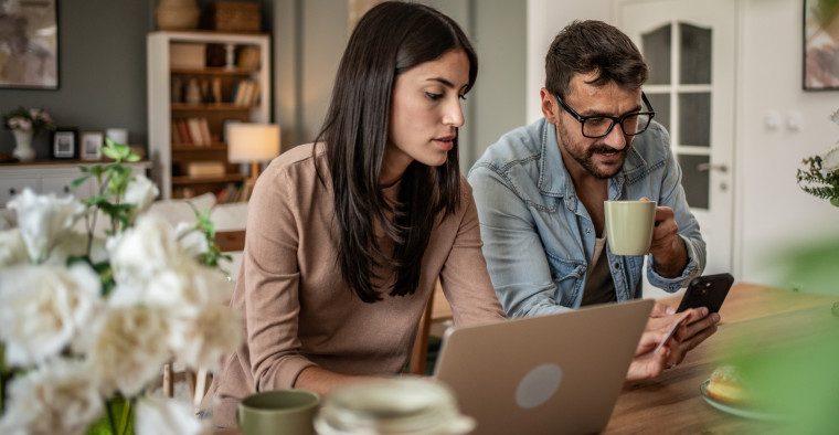 A couple sitting in a kitchen online shopping on a phone and a laptop