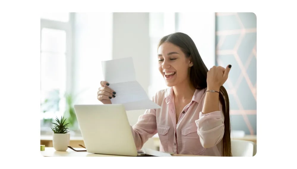 A young woman excitedly reading a letter while working on a laptop, representing career development or good news.png