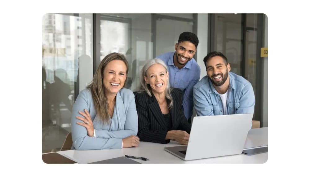 A cheerful group of professionals gathered around a laptop, smiling and engaging in a collaborative office environment.png