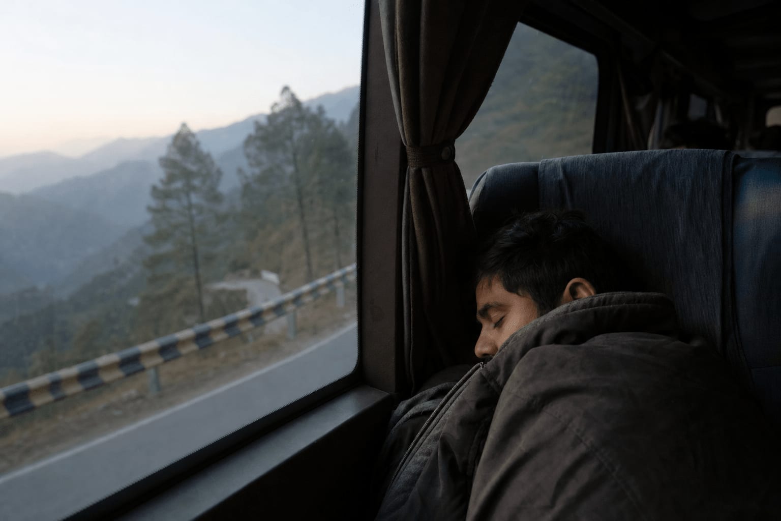 Veetrips Window seat on an overnight bus at dawn, pine-covered mountain road outside and a passenger asleep