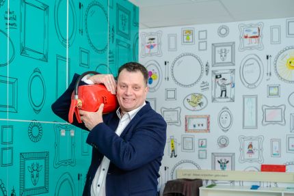 vrolijke foto van mario die met een brandblusser op zijn schouder in een kamer met gezellig behang van getekende fotolijsten staat