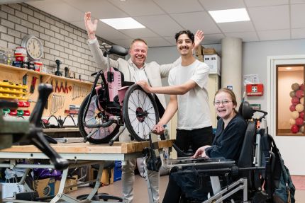 directeur bas staat vrolijk met zijn armen omhoog in de werkplaats op school achter leerling mo die wekrt aan een rolstoel en anne-louise die vrolijk lach vanuit haar rolstoel