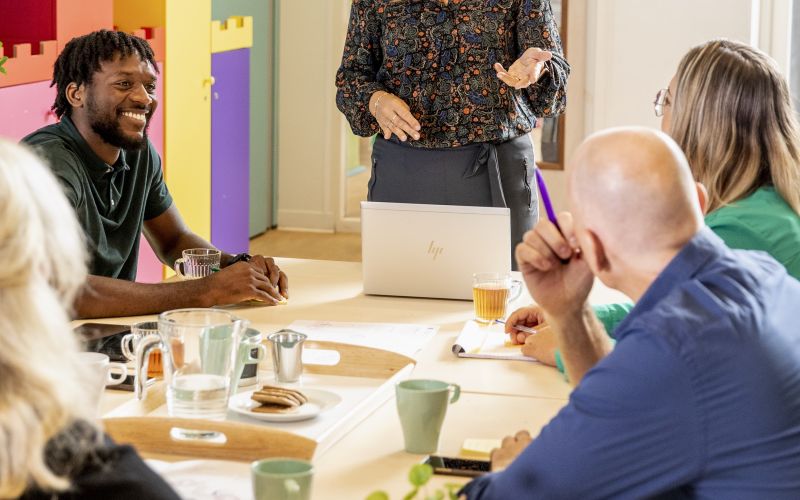 Vrouw spreekt groep collega's toe in kleurrijke klas met tekening vragende collega's