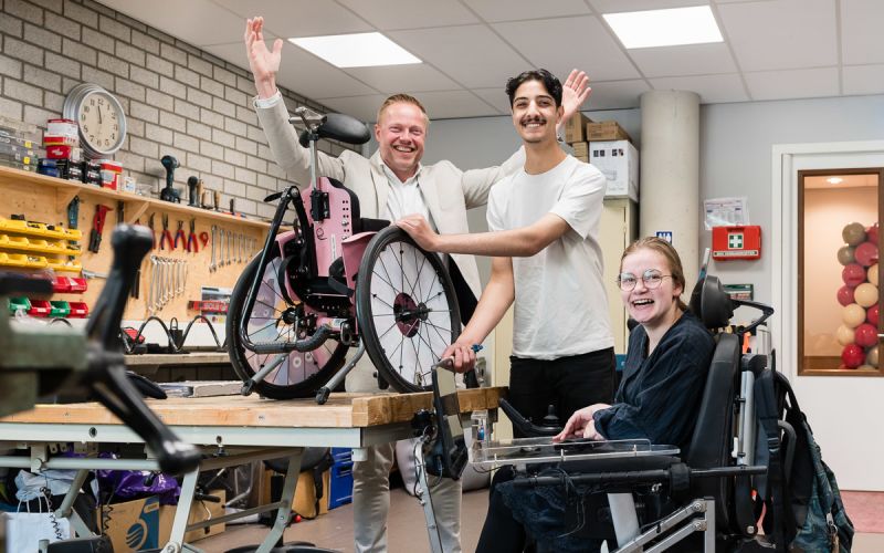 directeur Bas staat vrolijk met zijn armen omhoog in de werkplaats op school achter leerling Mo die wekrt aan een rolstoel en Anne-Louise die vrolijk lach vanuit haar rolstoel