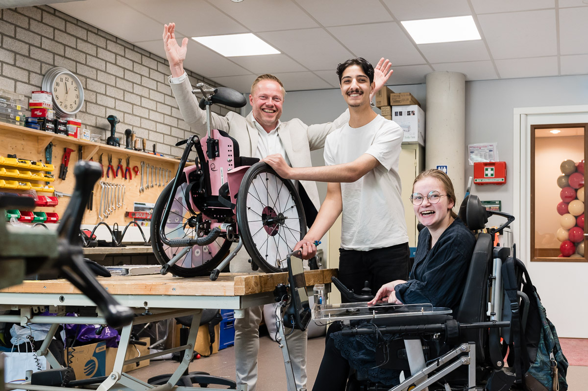 directeur bas staat vrolijk met zijn armen omhoog in de werkplaats op school achter leerling mo die wekrt aan een rolstoel en anne-louise die vrolijk lach vanuit haar rolstoel