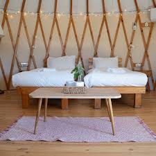 A cozy interior of a yurt featuring two twin beds, a wooden table, and a decorative rug. Soft lighting adds warmth to the space.