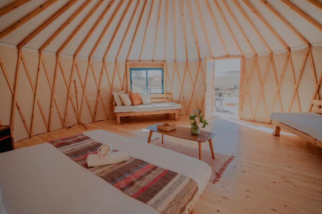 A cozy interior of a yurt featuring wooden beams, two beds, a small table, and a window with a view. Natural light fills the space.