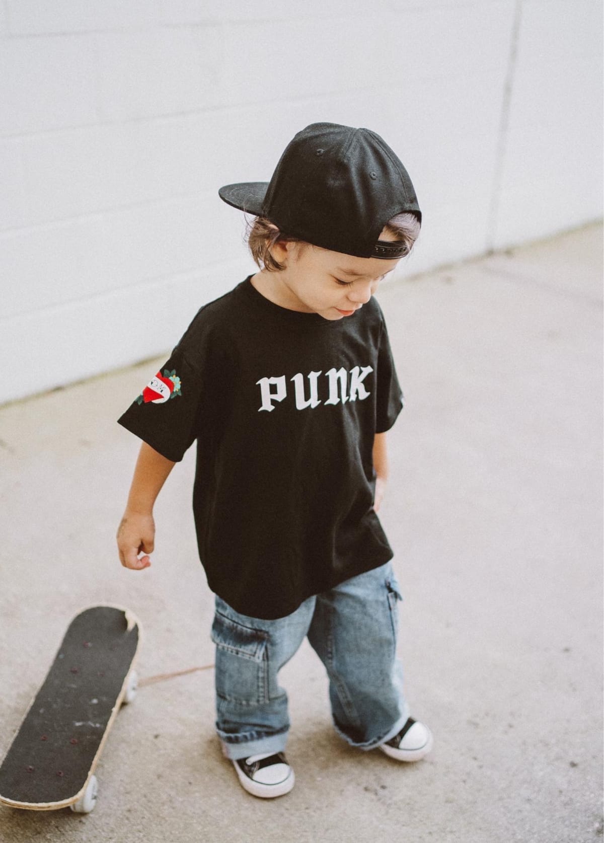 Child wearing a black PUNK T-shirt and backwards cap beside a skateboard