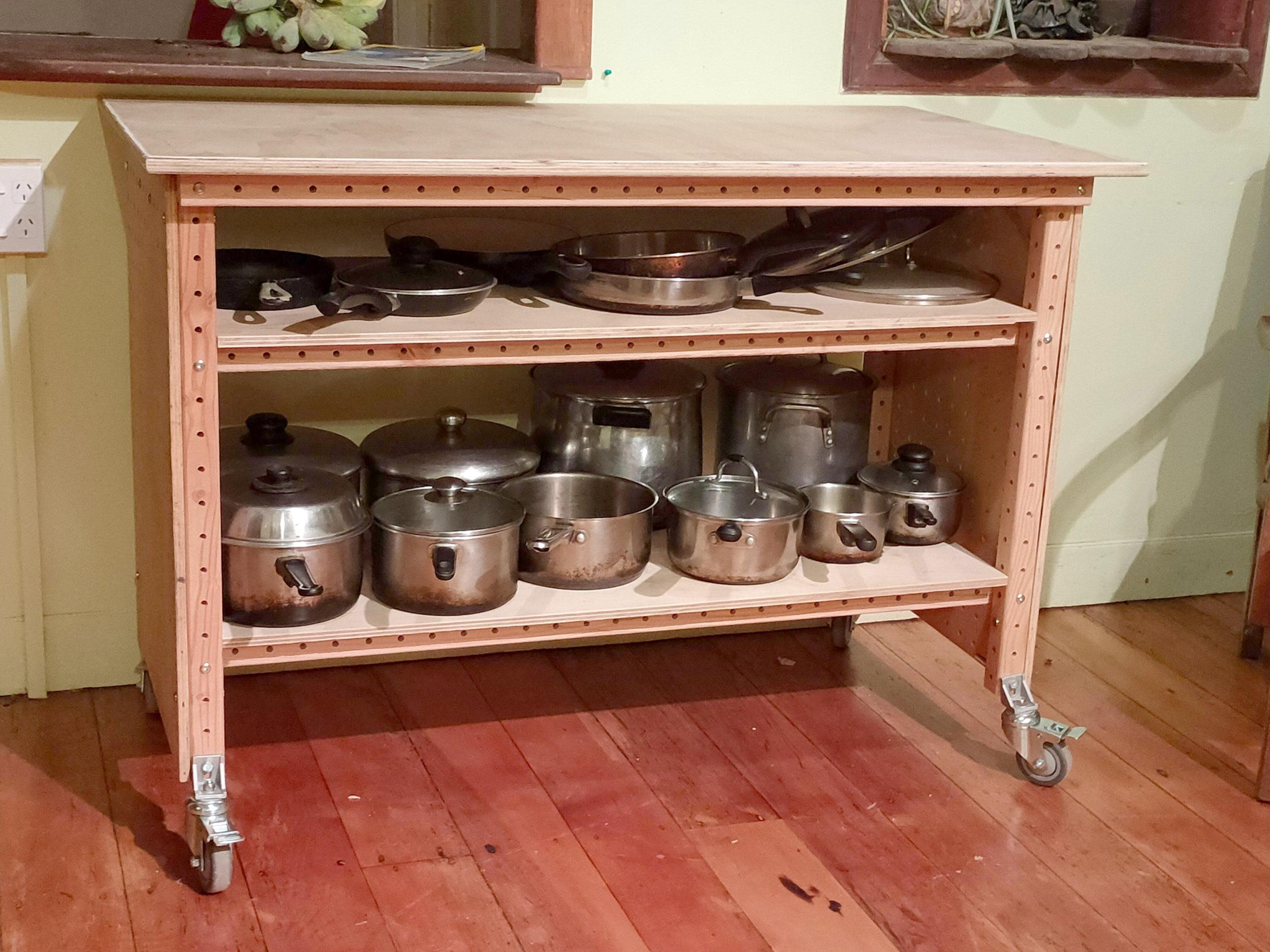 A wooden rolling cart with a smooth tabletop and two shelves underneath. The shelves store a variety of metal pots, pans, and lids. The cart has caster wheels and is set on a hardwood floor, with a windowed kitchen wall in the background.