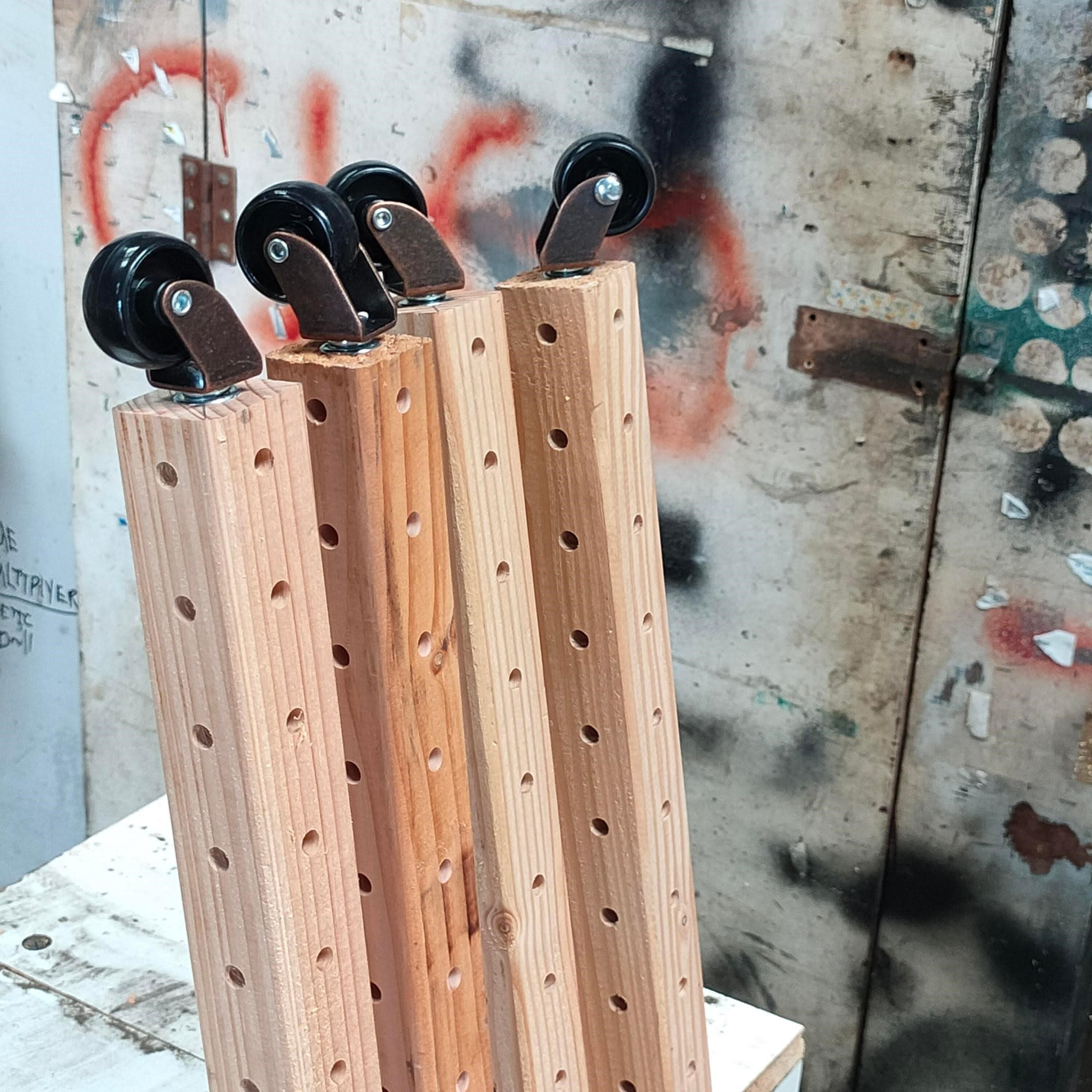 Four vertical wooden grid beams with caster wheels attached at the top, ready for mobility. The beams have evenly spaced perforations and are standing upright in front of a textured wall with spray paint marks.