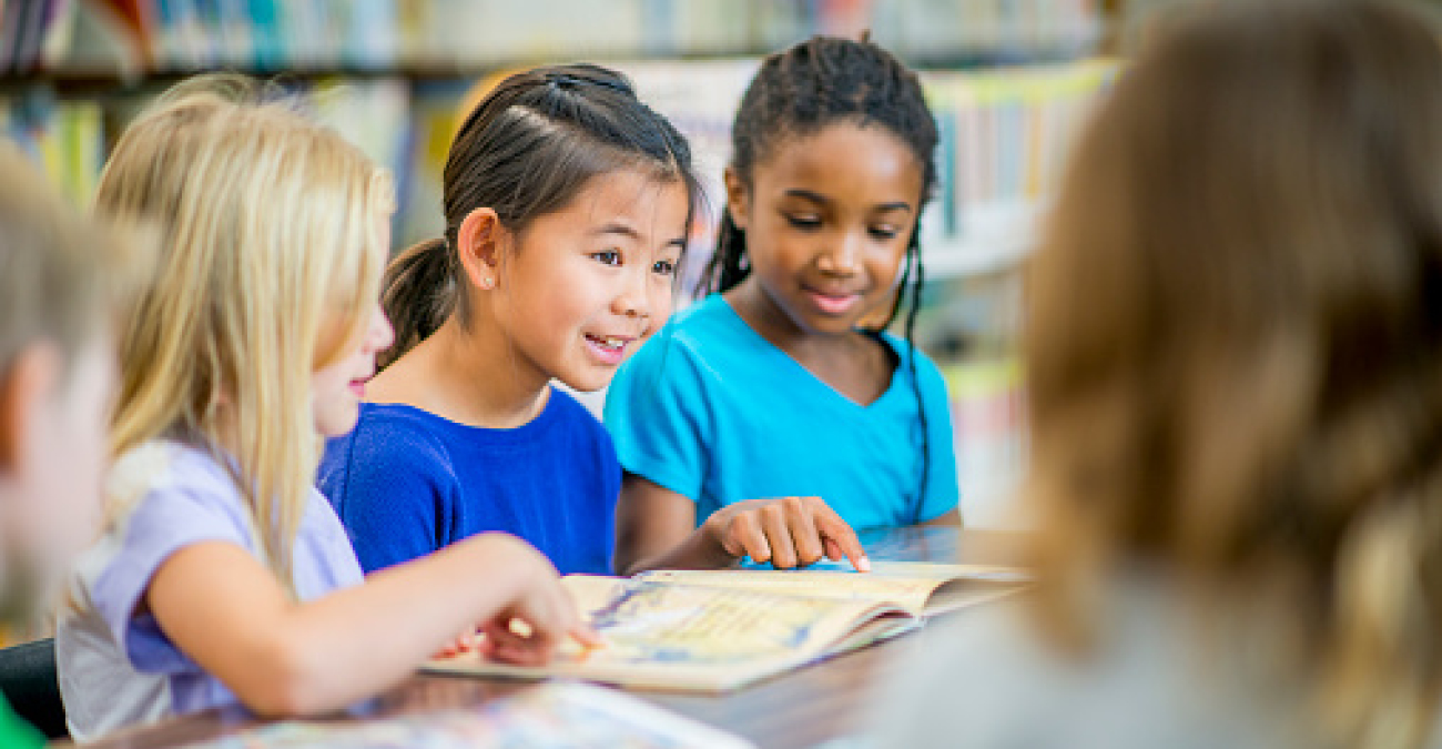 Three little girls read a book in a library. Two of the girls point to the contents of the book.