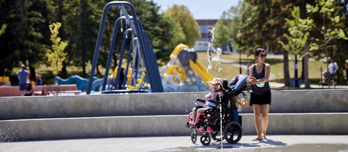 Enfant en fauteuil roulant et personne accompagnatrice près d’un jeu d’eau interactif, dans un parc muni d’aires de jeux.