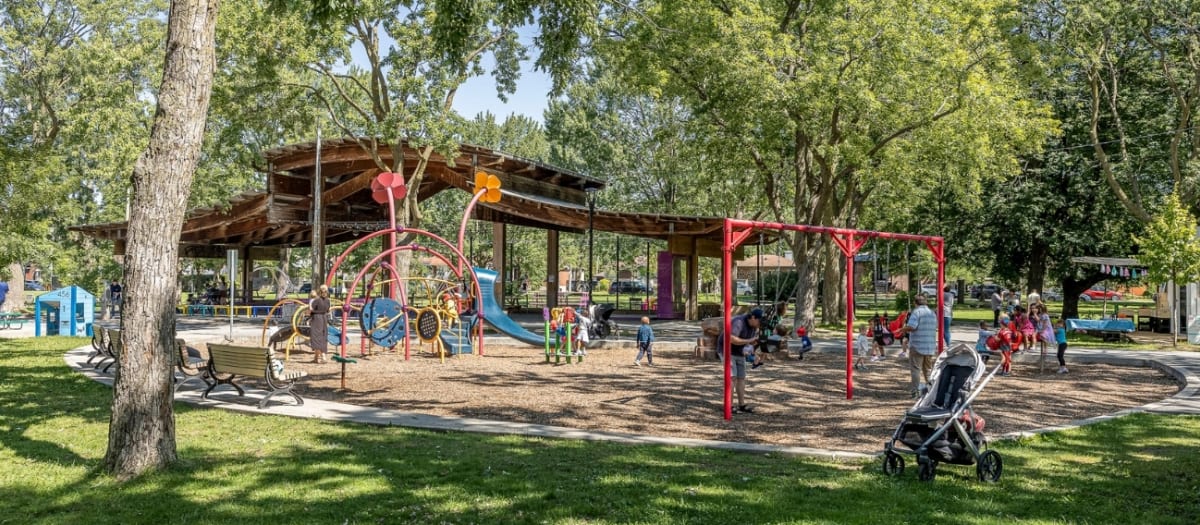 Photo d'une aire de jeux avec des enfants jouant dans les modules et les balançoires. En arrière-plan, on aperçoit un abri avec des terrains de boules.