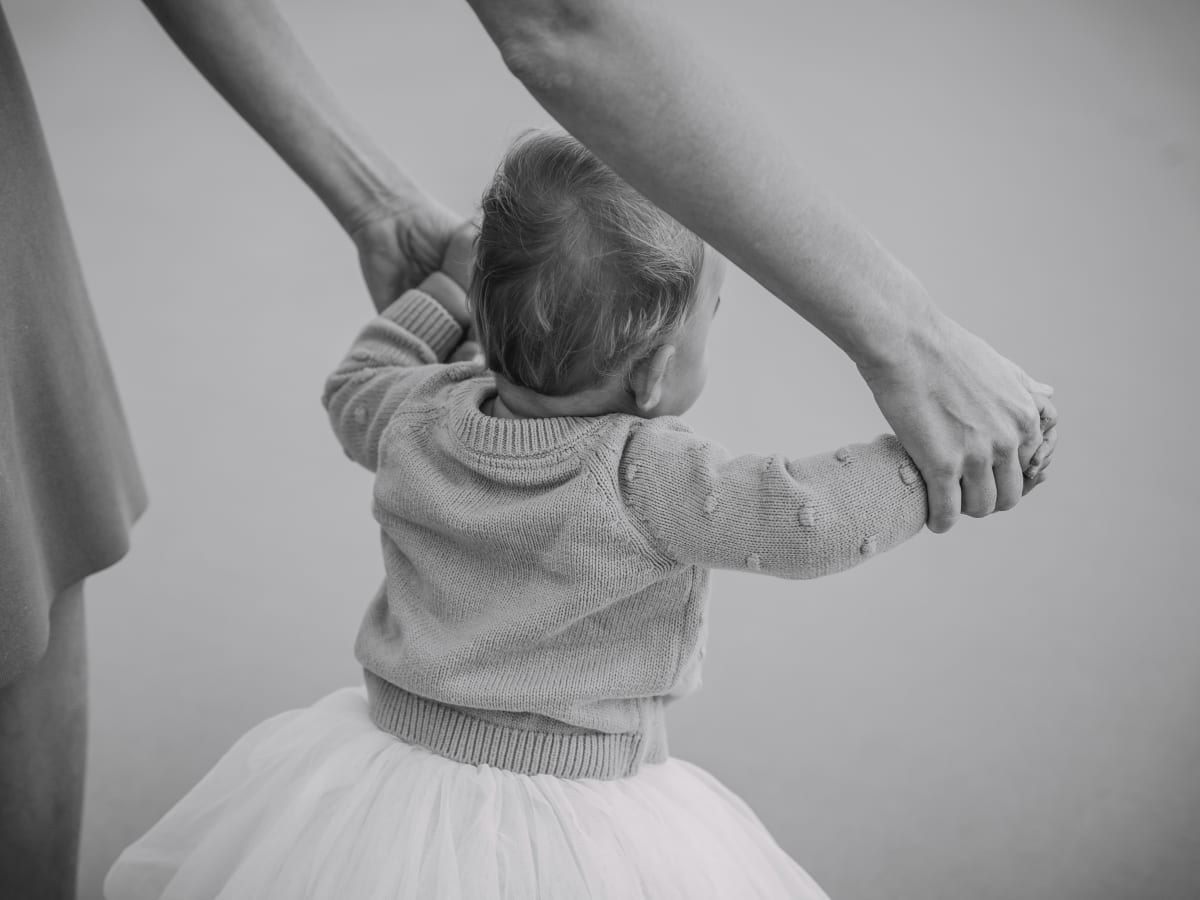 Photo en noir et blanc d'un poupon qui porte une veste et un tutu qui tient les mains d'un adulte