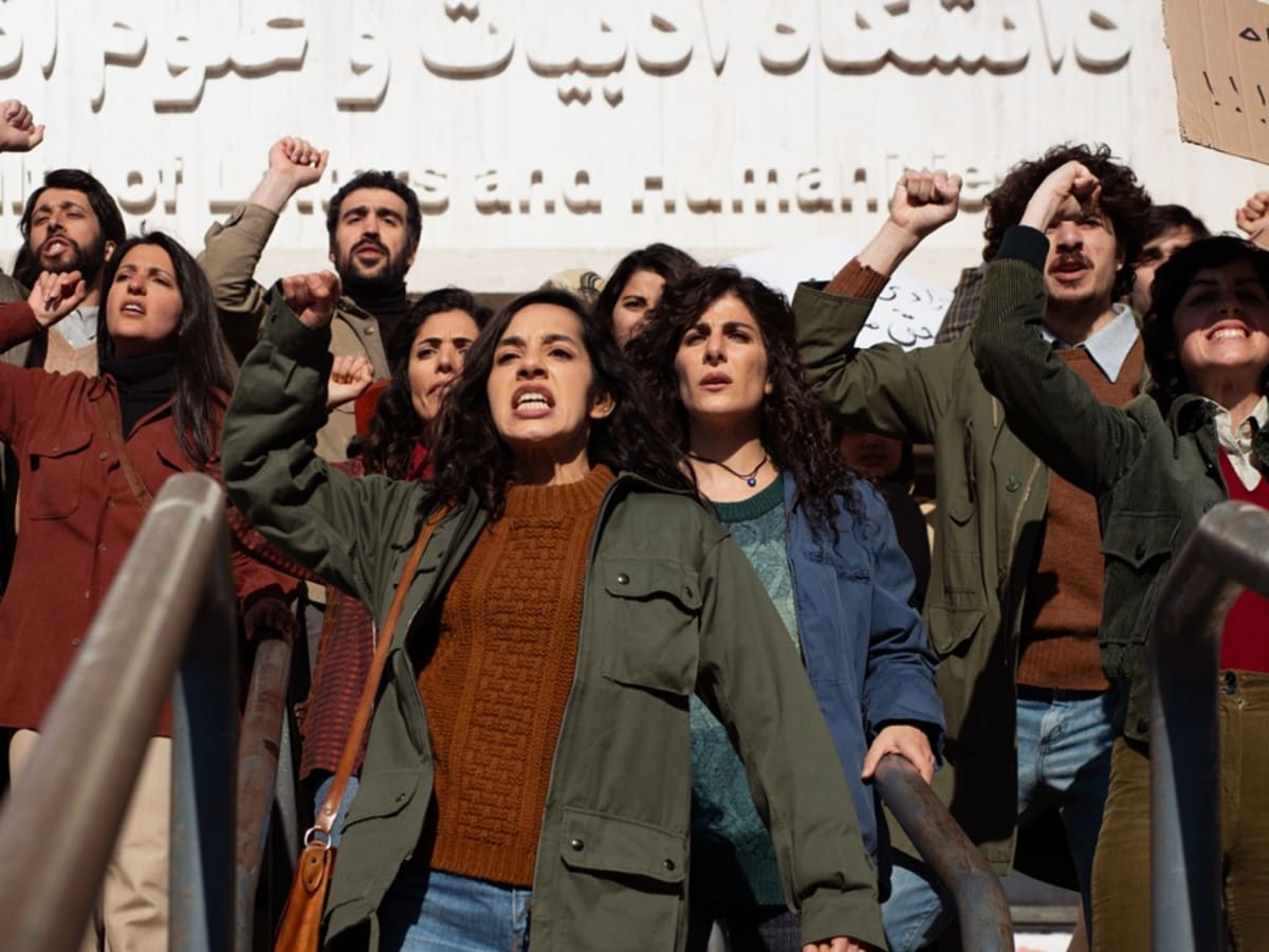 La photo montre un groupe de femmes qui manifestent, poing levé, devant un mur blanc avec des inscriptions en arabe. un