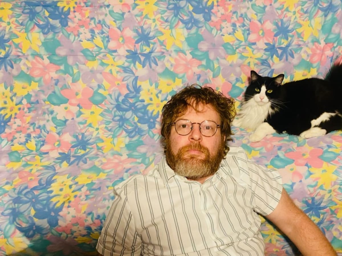 Un homme barbu portant des lunettes et un chandail à rayures se tient aux côtés d’un chat noir et blanc. L’ensemble est mis en valeur par un fond floral aux nuances de bleu, de jaune et de rose.