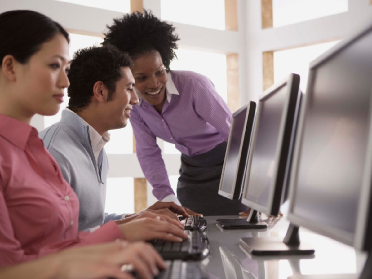 Des personnes à l'ordinateur dans une salle de formation informatique avec une personne qui les accompagne dans l’utilisation des postes de travail.
