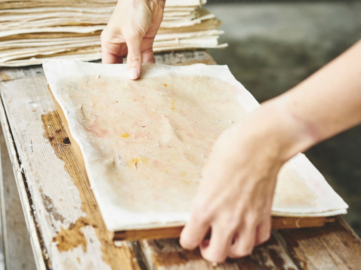 Des mains manipulent un tamis rectangulaire pour créer une feuille de papier artisanale.