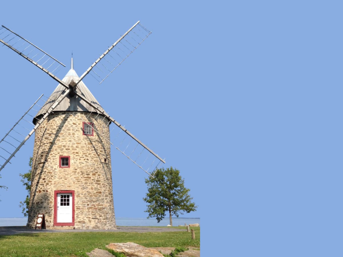 Un moulin à vent de pierre se dresse près du fleuve sous un ciel bleu dégagé, entouré de quelques arbres et d’une pelouse verdoyante. L’image évoque le patrimoine et la vie quotidienne à l’époque de la Nouvelle-France.