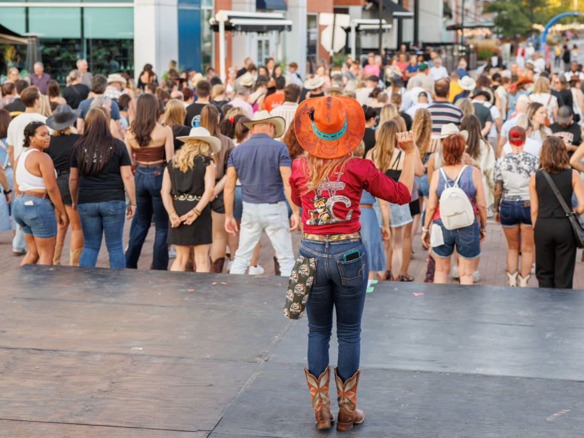 Une personne qui porte un chapeau de style western se tient sur une scène devant une foule rassemblée dans une rue animée lors d’un événement extérieur.