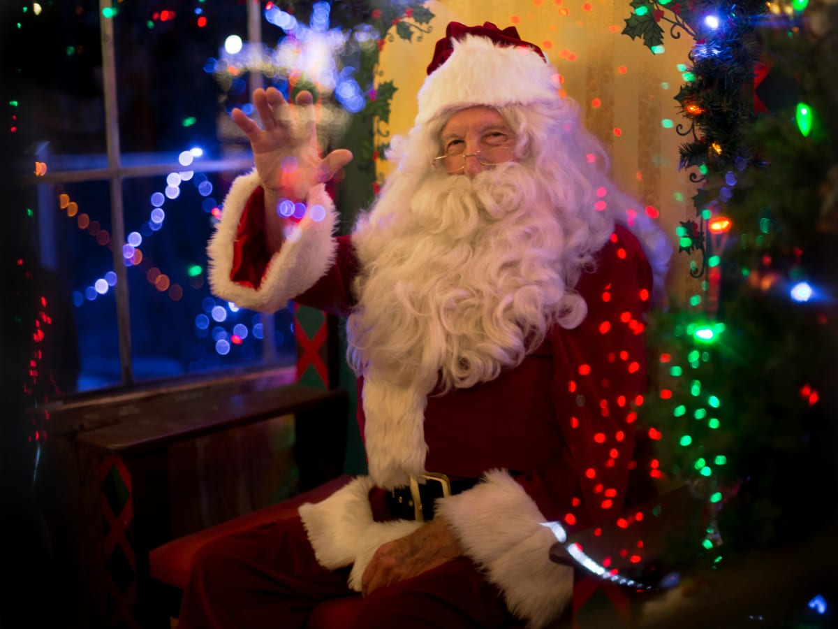 Photo du Père Noël avec une grande barbe blanche portant un manteau et patalon rouges et blanches assis à côté d'une sapin avec des lumières colorées