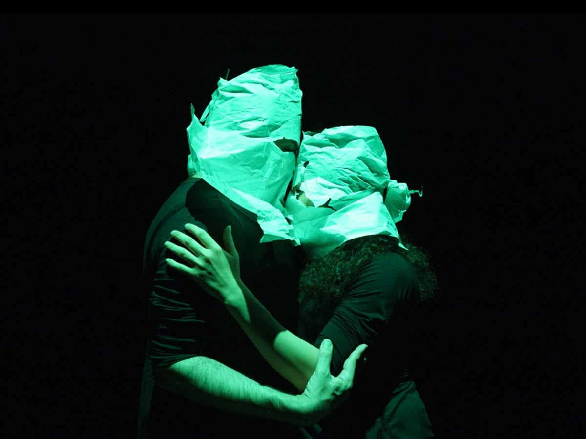 Couple de danseurs cagoulés et enlacés sur scène, avec arrière plan très sombre.