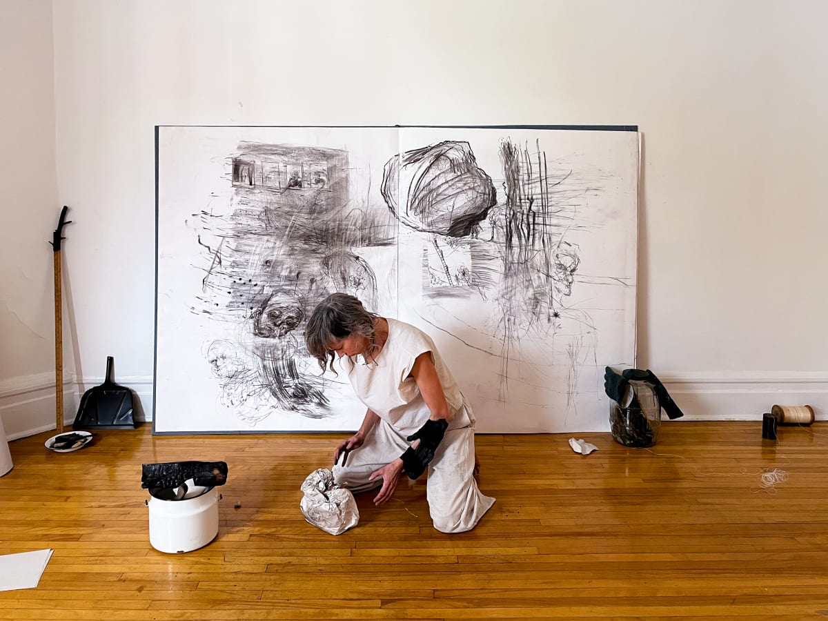 Personne vêtue de blanc agenouillée sur un plancher de bois, manipulant un objet froissé devant une grande œuvre monochrome composée de dessins au fusain. Divers outils et matériaux artistiques sont disposés autour de l’espace de travail.