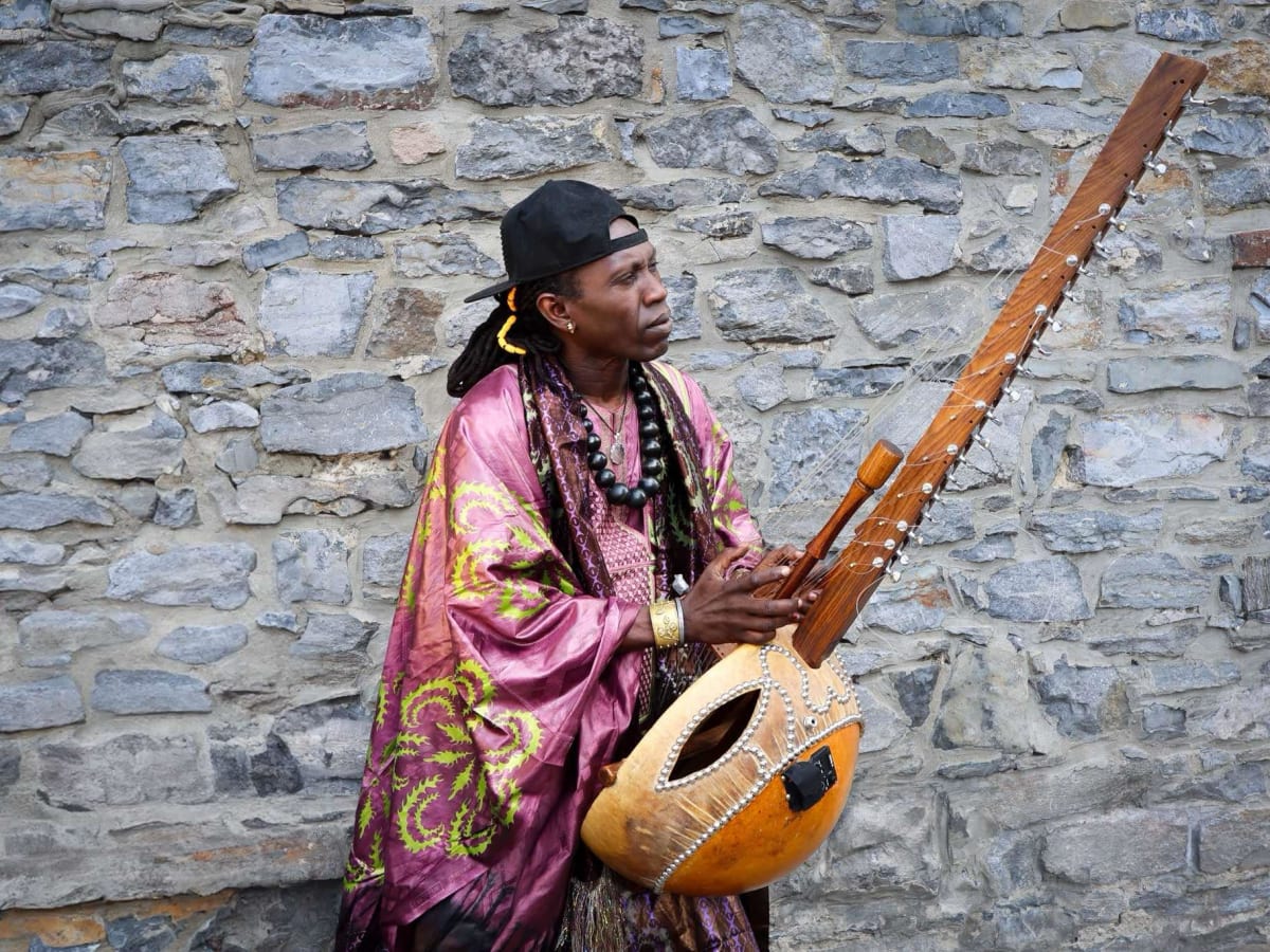 La photo représente le musicien tenant sa kora devant un mur gris