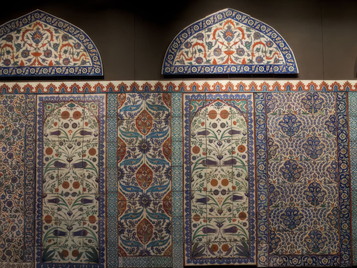 Photographie d'un mur de mosaïques islamiques formé de motifs géométriques dans les teintes de bleu, de beige et d'orangé. La mosaïque est composée de 6 panneaux différents (sauf 2), surmonté d'une frise à dominance beige et de trois ogives.