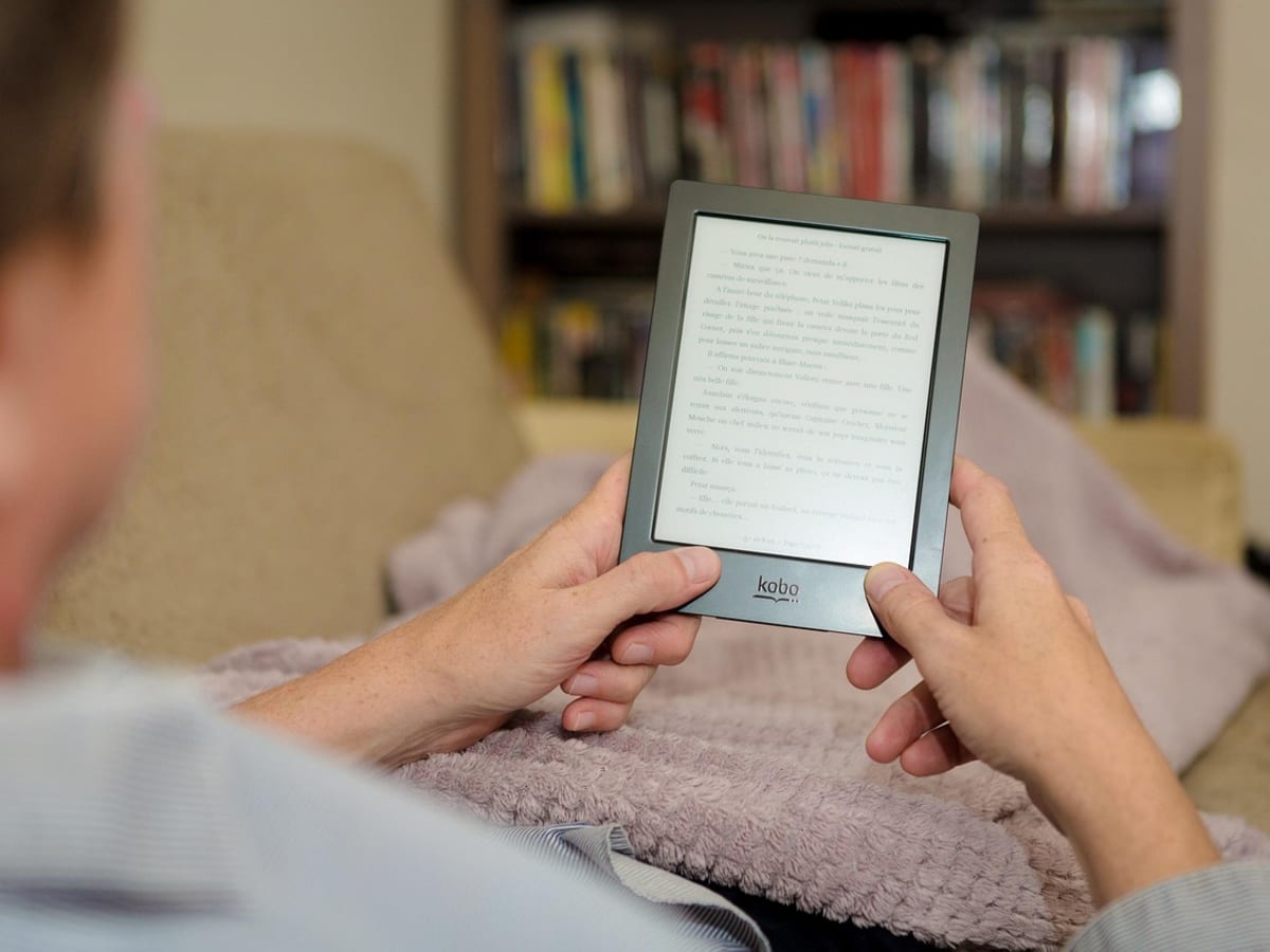 Photo d'une personne assise, avec une couverture sur les jambes qui tient entre ses deux mains un tablette de marque Kobo. À l'arrière plan, on voit une bibliothèque.