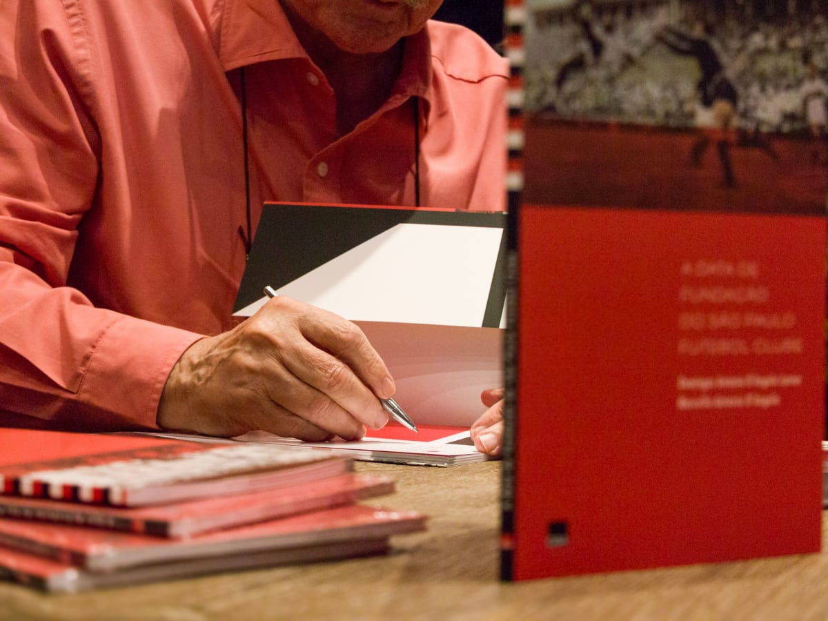 Une personne assise à une table signe un livre lors d’une séance de dédicace. Plusieurs exemplaires du livre sont disposés sur la table, dont un ouvert au premier plan.