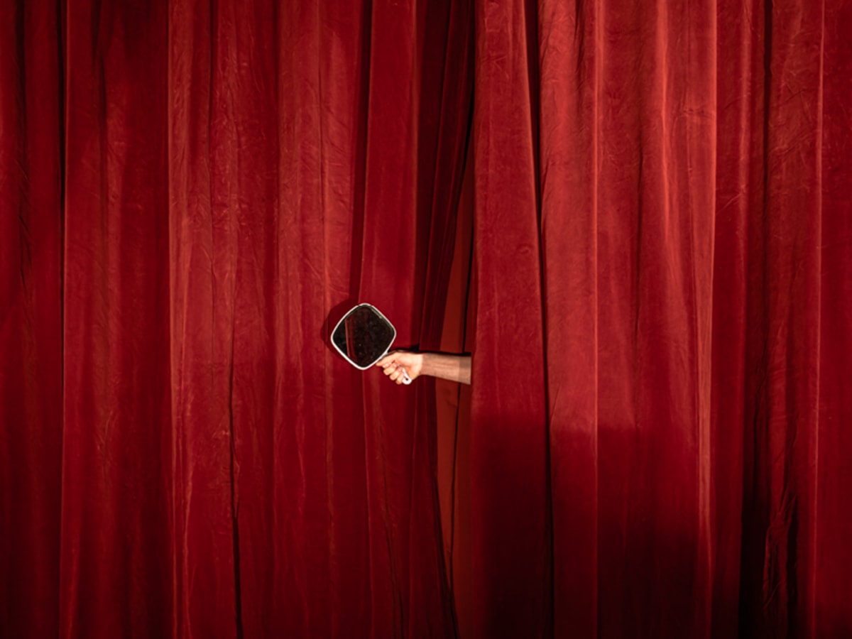 Une main tenant un petit miroir sort d'un grand rideau de théâtre rouge.