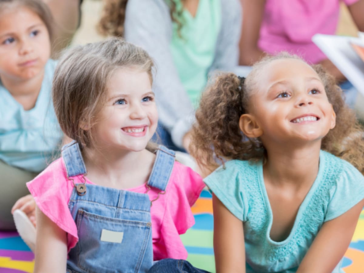 Deux jeunes enfants souriantes regardent quelqu'un qui lit un livre.