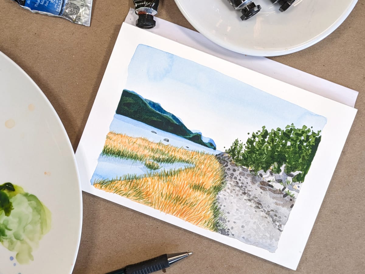 Une photo d'une aquarelle représentant un paysage, entouré de tube de peinture aquarelle