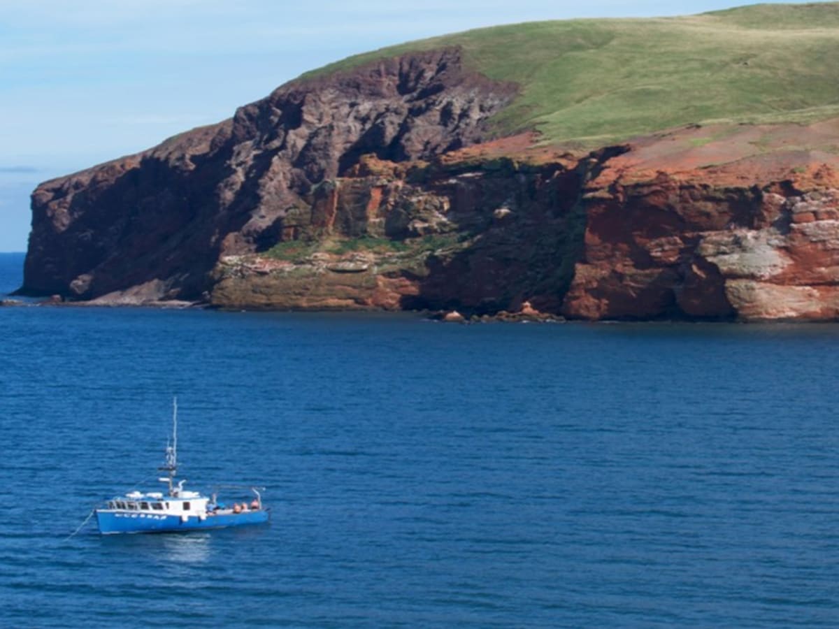 paysage d'un bateau blanc navigant sur une mer calme et bleue. Le ciel est bleu strié de vaporeux nuages. Une grosse falaise verdoyante occupe l'arrière-plan.