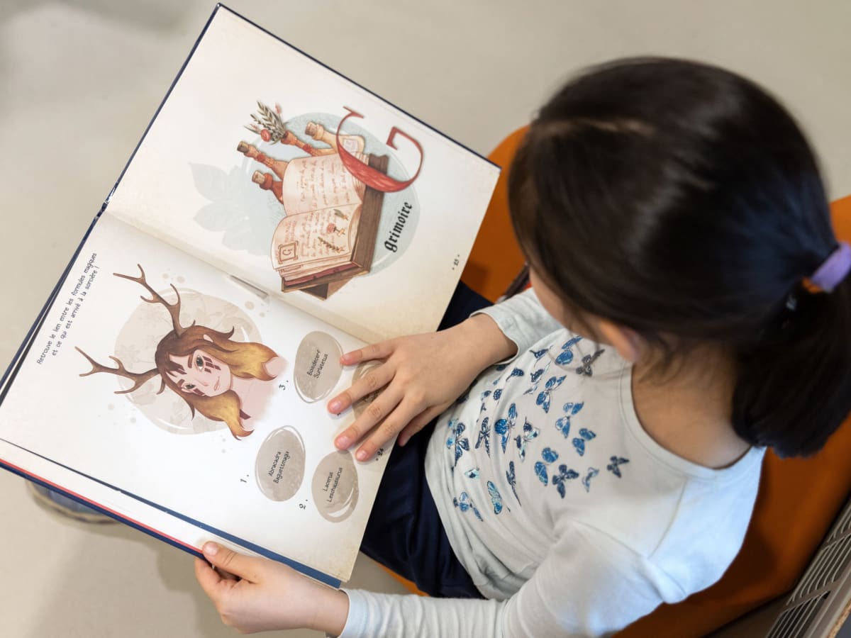 Une enfant assise qui lit un grand livre illustré.