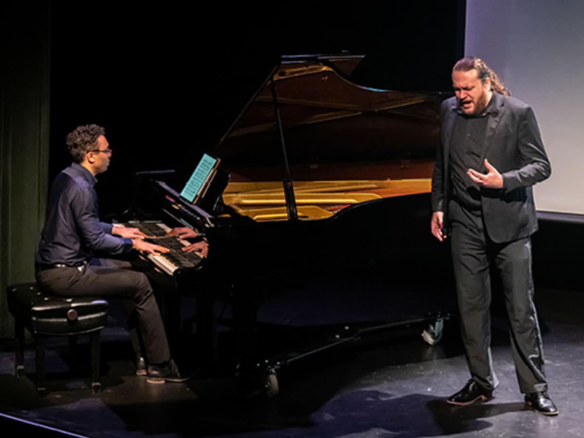 Sur scène, un homme en costard noir chante accompagné d'un pianiste.