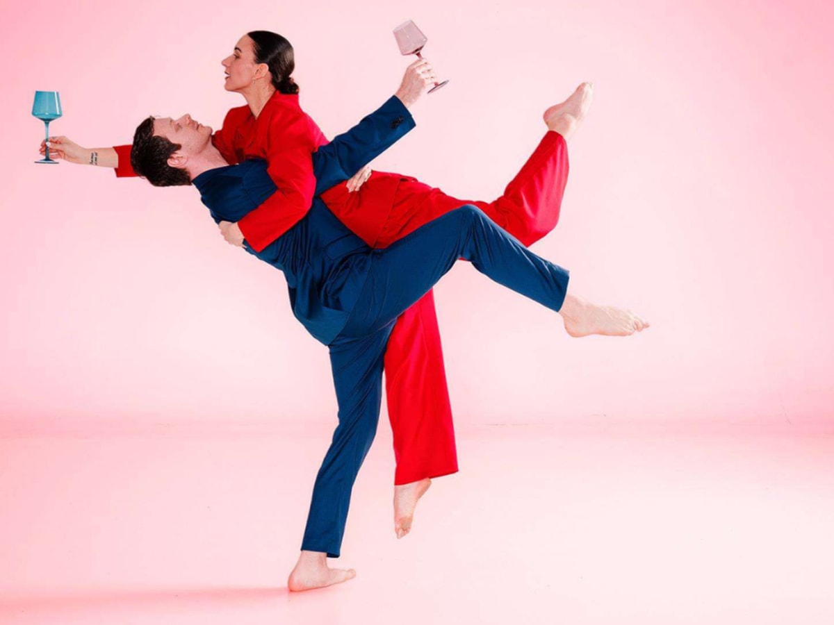 Deux danseurs se prennent dans leurs bras les pieds dans les airs, tenant chacun un verre de vin. Ils sont sur fond rose.