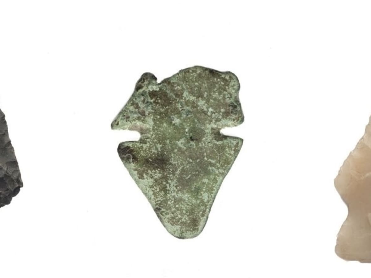Trois pointes de projectile préhistorique de formes triangulaires, disposées côte à côte sur un fond blanc : une pointe en pierre sombre à gauche, une pointe de cuivre verdâtre au centre et une pointe en pierre claire à droite.