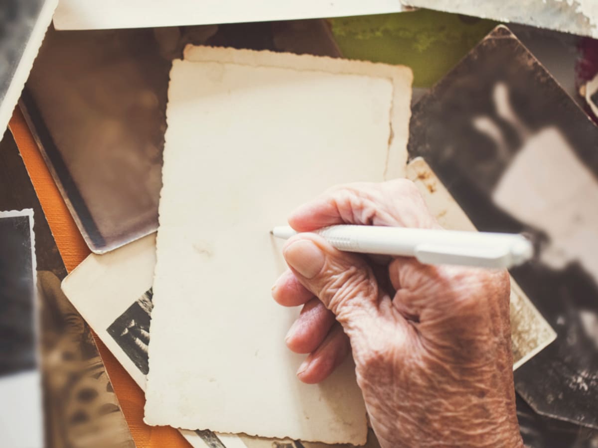 Une main tient un stylo au-dessus d’une feuille vierge, entourée de photographies anciennes éparpillées. L’image évoque l’écriture de souvenirs et le partage de récits de vie à partir de photos du passé.