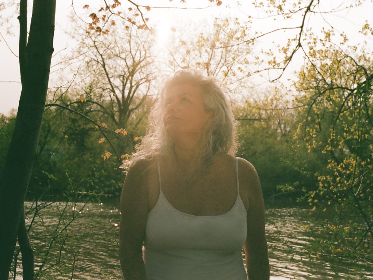 L'artiste Monica Freire devant un lac et une foret, les couleurs sont jaune et verte.
