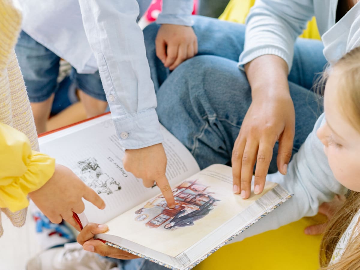 Des enfants sont rassemblés autour d’un livre ouvert et pointent les illustrations pendant une activité de lecture.