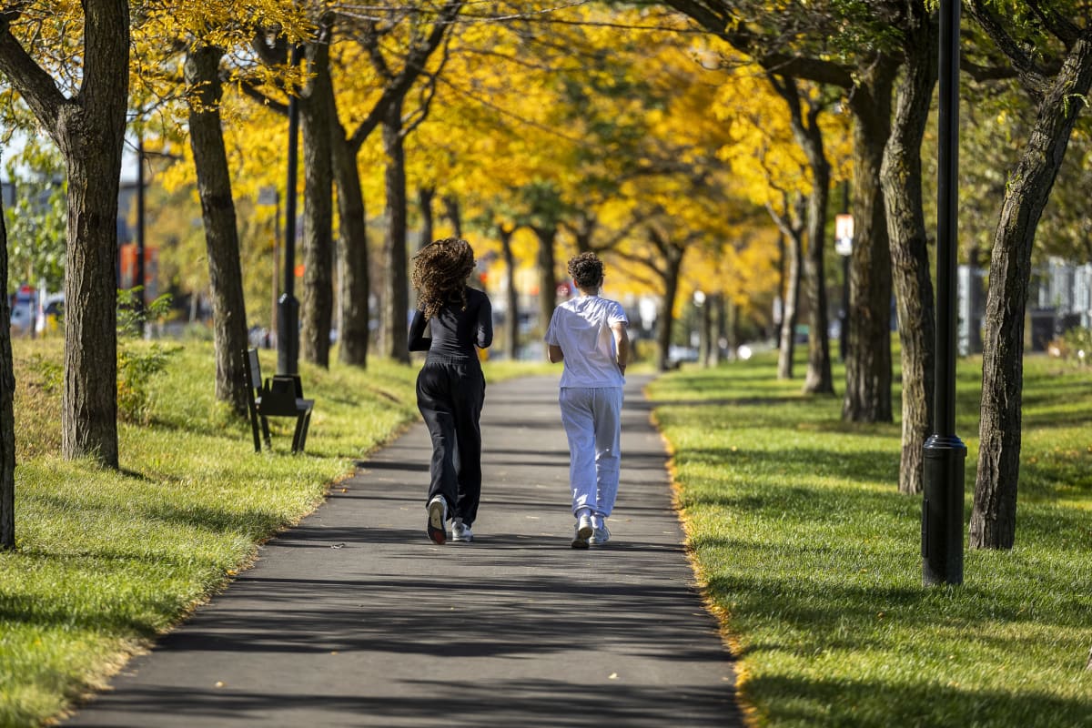 Photo de deux femmes courant sur un sentier asphalté bordé de gazon et d'arbres au feuillage automnal.