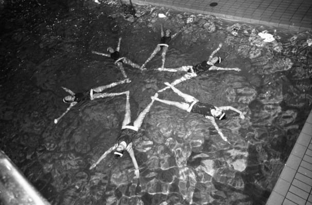 Photo en noir et blanc montrant des nageuses de natation artistique allongées dans l'eau d'une piscine, en train de former une étoile avec leurs jambes unies