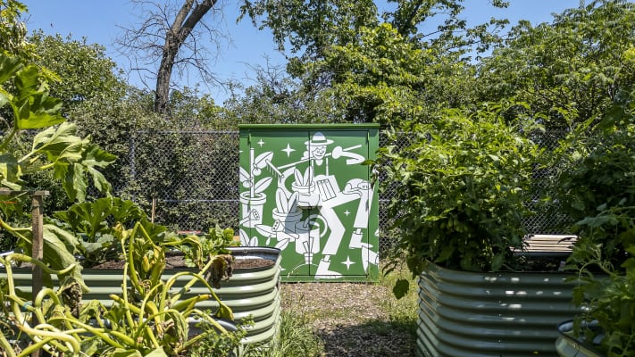 Photo d'une grande armoire en métal au milieu d'un jardin luxuriant. L'armoire est de couleur verte et affiche une illustration de personnage en train de jardiner.