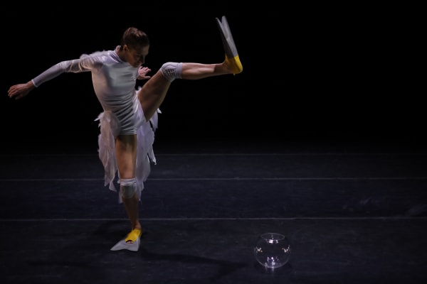 Une actrice avec des palmes lève exécute un mouvement de danse alors que se trouve un bocal en verre sur le sol.