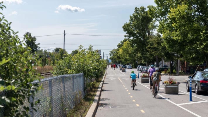 Une famille circulant à vélo sur la portion du REV située sur l’avenue de Souligny, entre la voie ferrée et la rue. Les vélos sont protégés par une zone tampon tracée sur le sol, des bollards et des blocs de béton fleuris à intervalles réguliers.