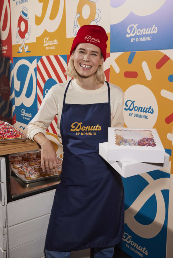 A woman wearing a branded hat and apron for a donut shop, with matching branded posters behind her
