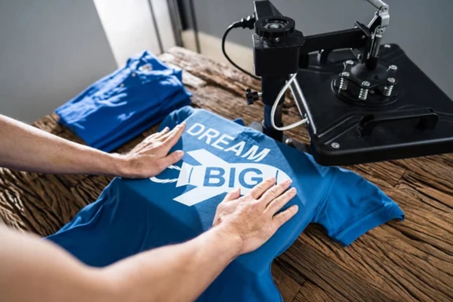 Hands positioning a blue T-shirt under a heat press, with a white &ldquo;dream big&rdquo; design on the fabric.