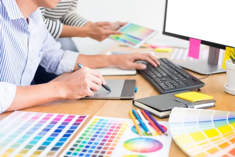 Designers working at a desk with a drawing tablet, computer and printed color swatches.